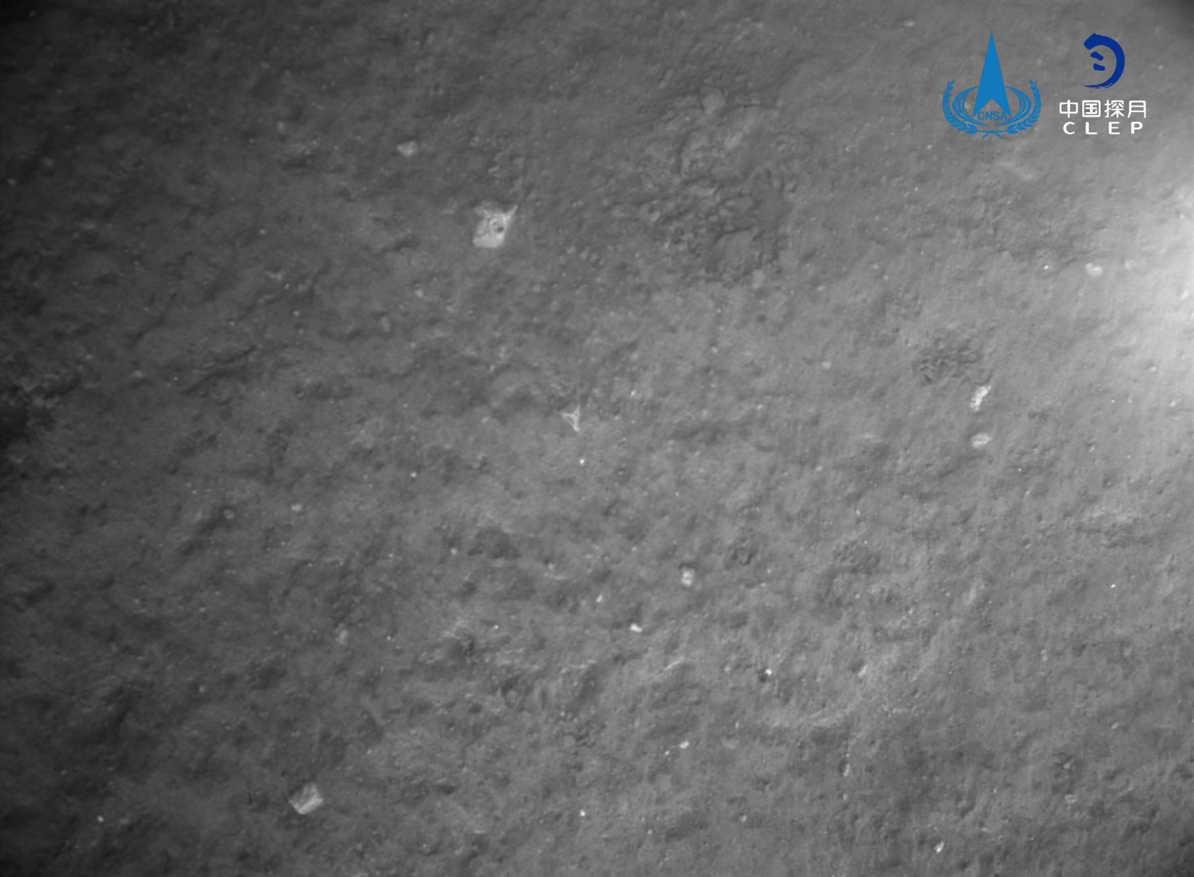 该图由降落相机在着陆器安全着陆后拍摄，图像显示着陆器底部相对平坦，分布有少量亮色石块。
